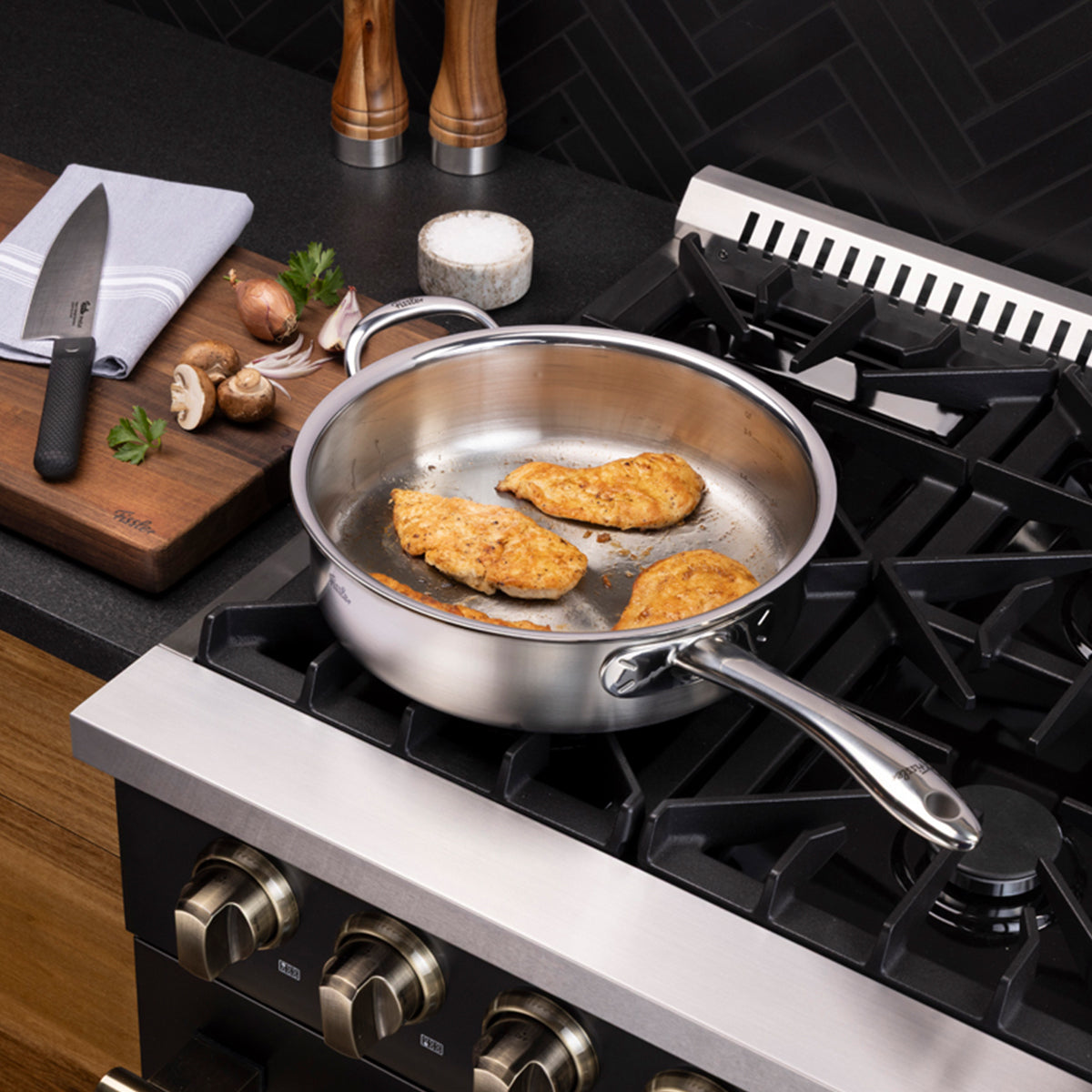 Drei Hähnchenteile werden fachmännisch im 3-teiligen Fissler M5 Pro-Ply®-Set mit Metalldeckel auf einem Gasherd zubereitet. Daneben liegen auf einem Schneidebrett ein Messer, frische Pilze, Knoblauch, Kräuter und ein Handtuch. Das hochwertige, induktionsgeeignete Fissler-Kochgeschirr – seit 1845 in Deutschland präzise gefertigt und bekannt für seine Langlebigkeit und sein zeitloses Design – hebt sich deutlich von der eleganten, modernen schwarzen Küchenrückwand ab.