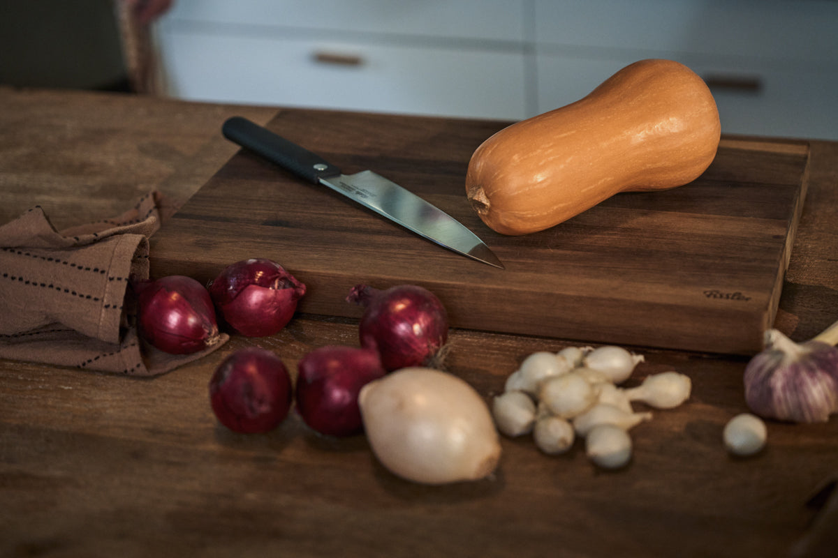 Auf einem Tisch liegen ein Butternusskürbis, Zwiebeln, Knoblauch und ein hochwertiges Messer auf einem hölzernen Fissler-Schneidebrett. Daneben liegt ein ordentlich gefaltetes Tuch – alles in einer einladenden Küche, die die Präzision, Langlebigkeit und das zeitlose Design der hochwertigen Fissler-Küchenutensilien widerspiegelt.