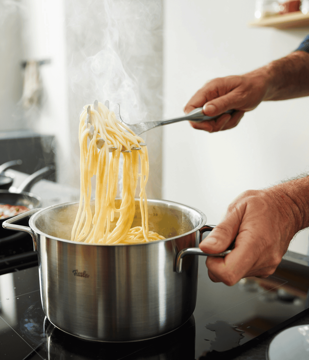 A home chef lifts spaghetti with tongs from a Fissler stainless steel pot, showcasing German precision and timeless design.